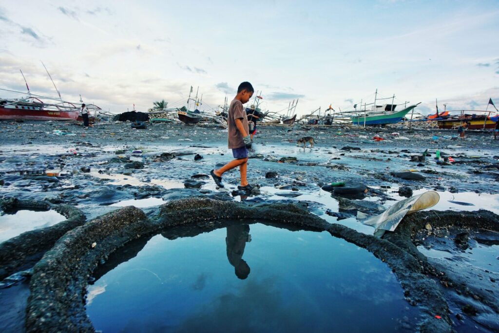 A child walks through a polluted beach with moored boats and debris, reflecting environmental issues.